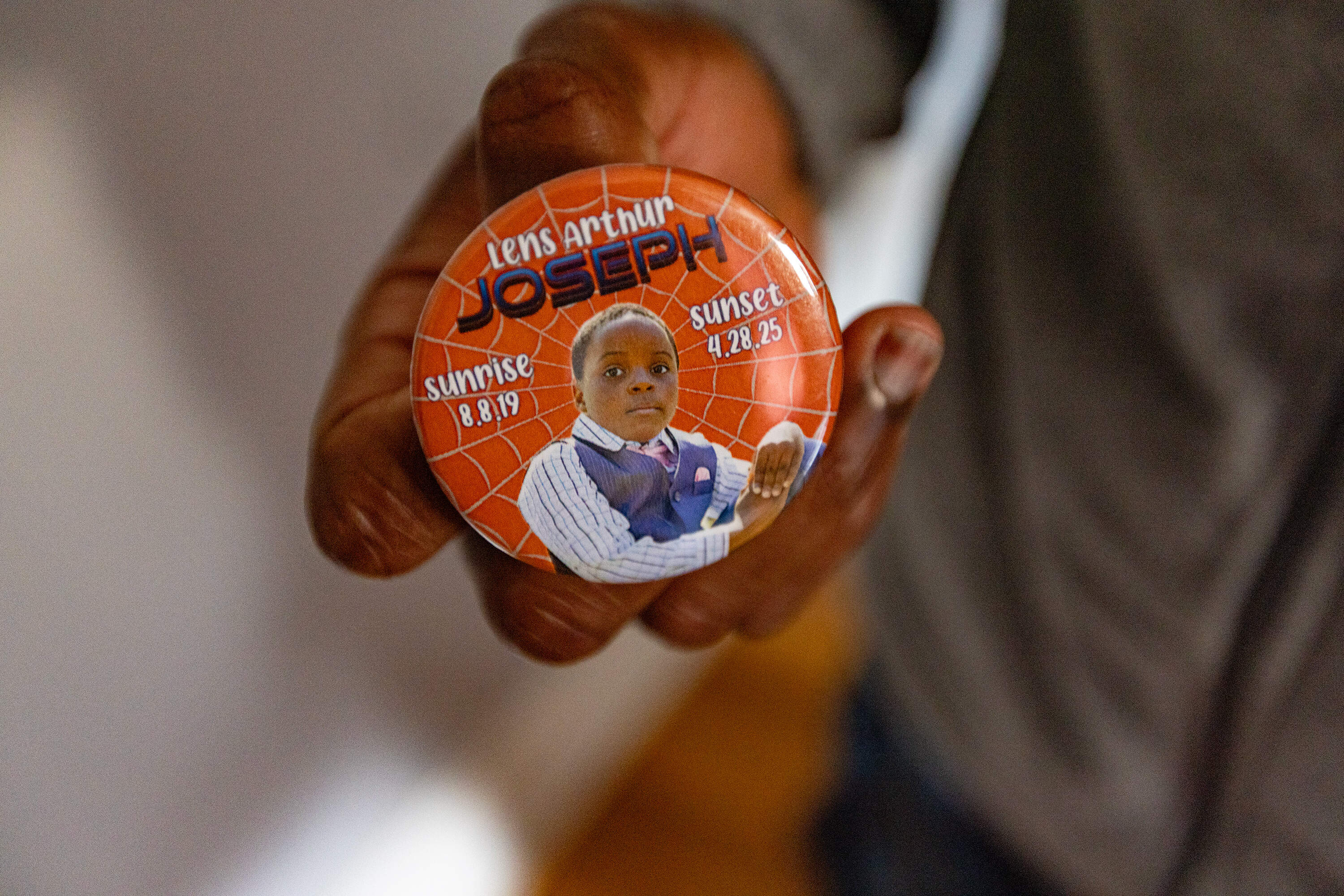 A man holds a button that has a photograph of a young child on it, and the words, “Lens Arthur Joseph. Sunrise 8.8.19. Sunset 4.28.25.”