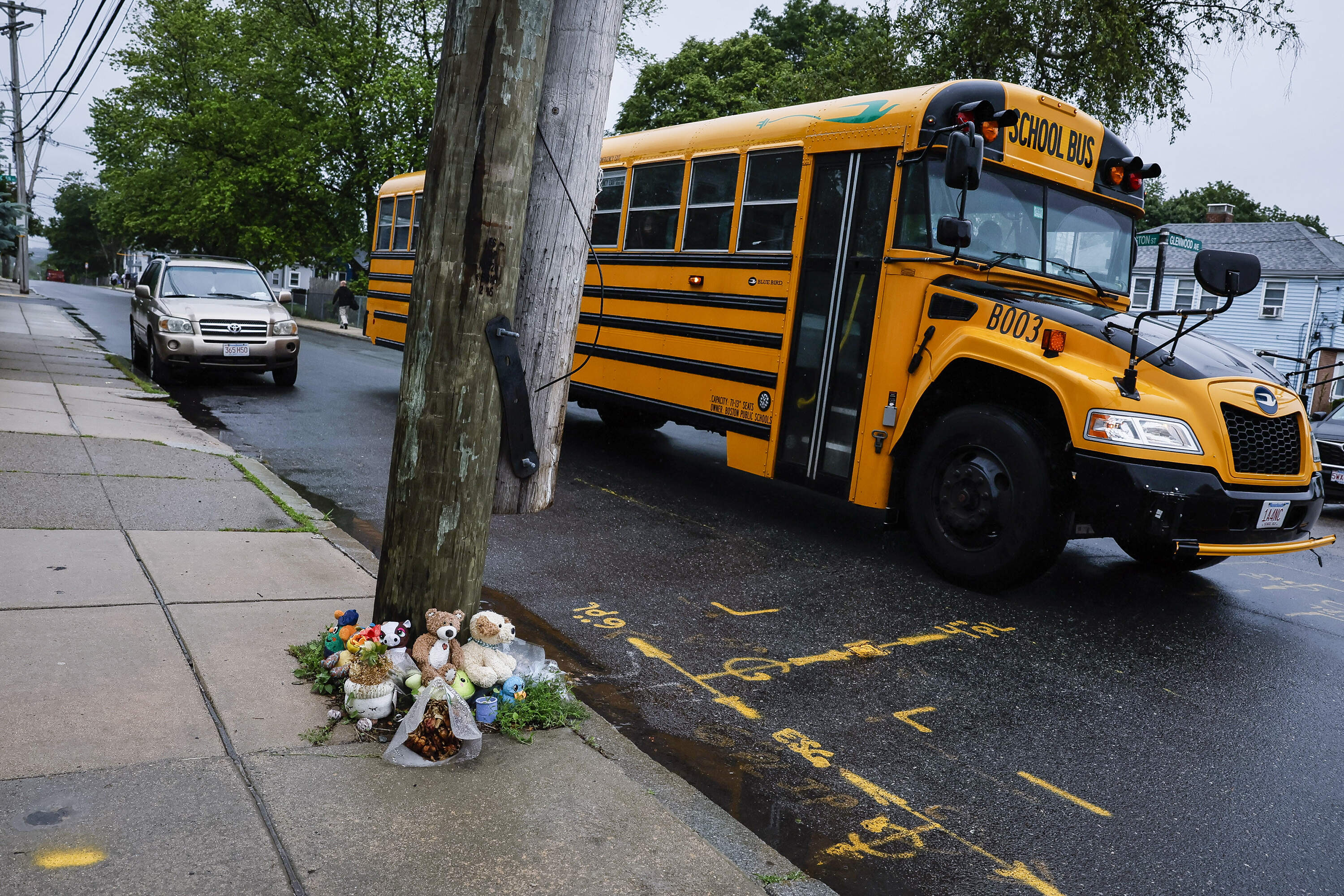 An orange school bus on a city street next to a sidewalk memorial made up of stuffed animals and flowers.
