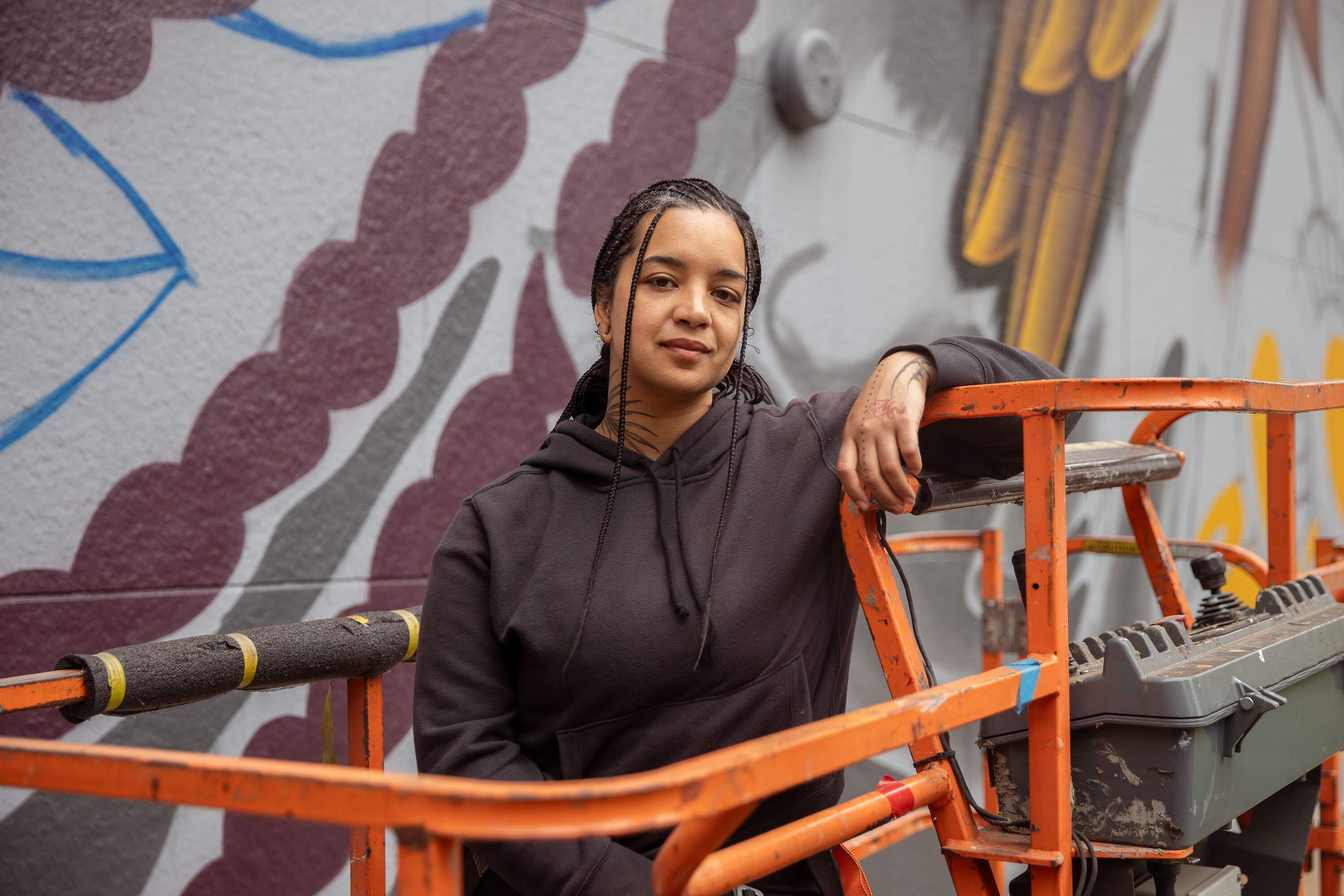 Lawrence-based artist and 2026 Dewey Square muralist Rixy poses in front of her in-progress Dewey Square mural on May 21. (Artemisia Luk/WBUR)