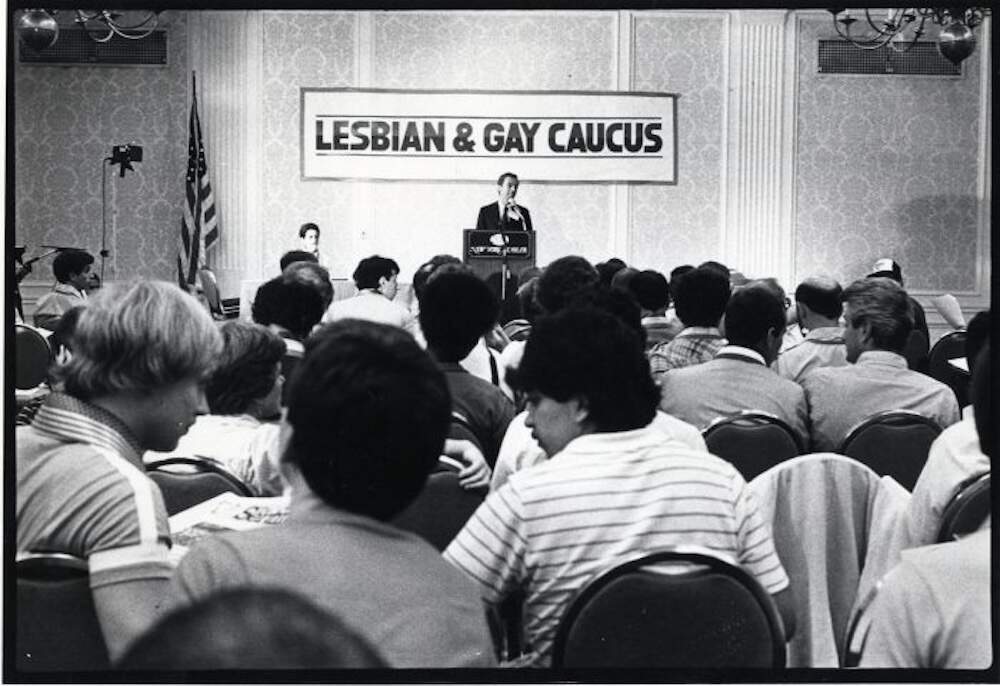 Tom Barstow, co-director of Gay Vote '80, addressing a large audience from a podium under a banner: "Lesbian and Gay Caucus." (Courtesy Queer History Boston)
