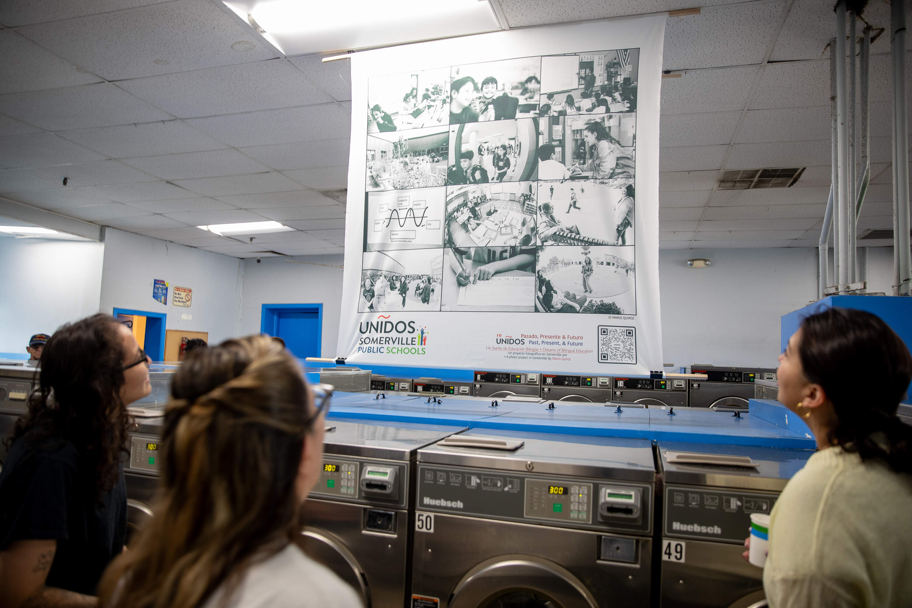 Melanny Motto, Alexandra Fuentes and Beatriz Thomas view Mario Quiroz’s photographs at City Line Laundromat during the opening reception on Wednesday, Sept. 16, 2026. Motto and Thomas are alumni of ESCS, where Quiroz works and captured the photographs. (Artemisia Luk/WBUR)