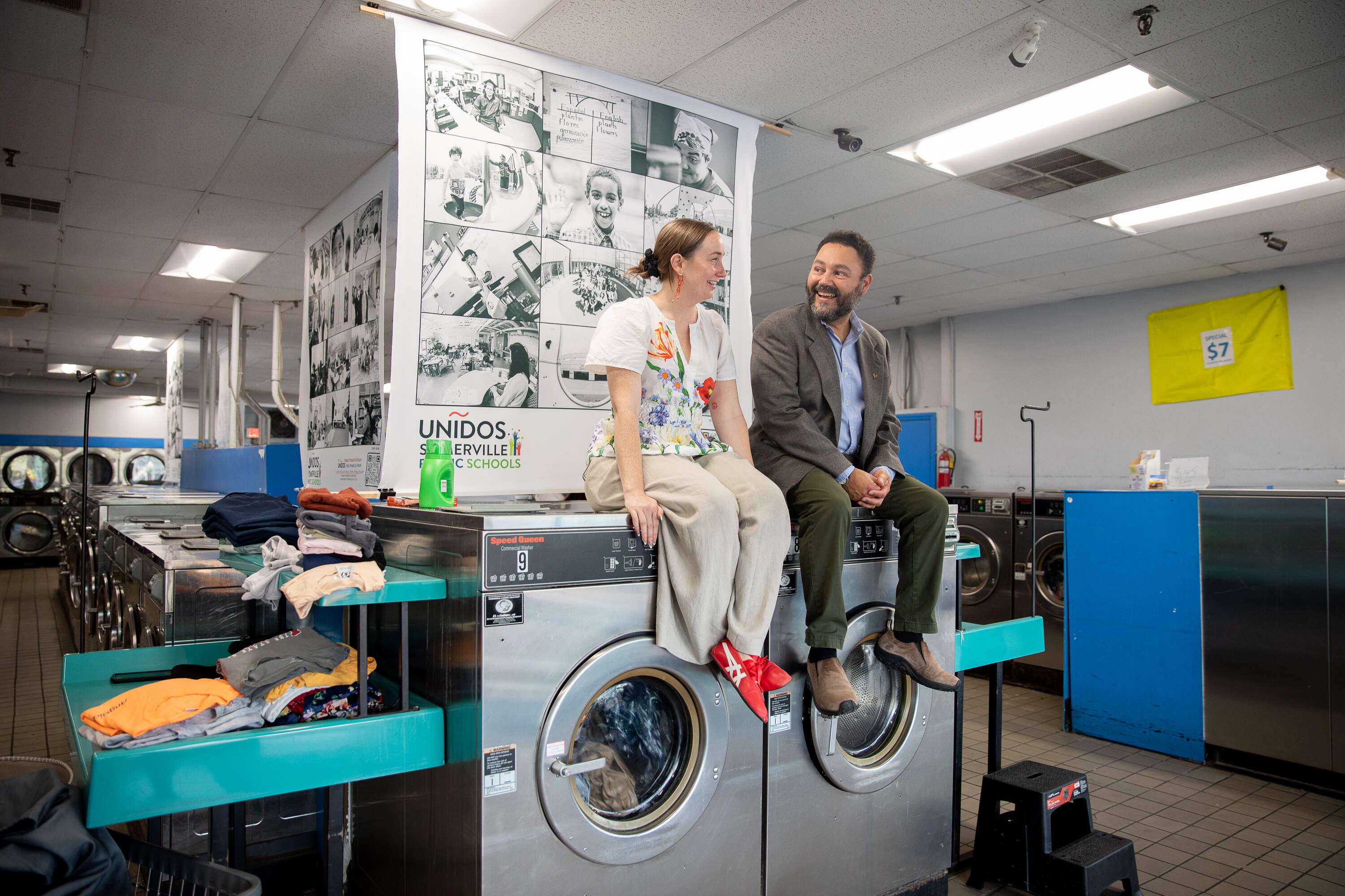 Unidos program specialist Analiese Reigstad and photographer Mario Quiroz pose in front of Quiroz’s photographs at City Line Laundromat. (Artemisia Luk/WBUR)